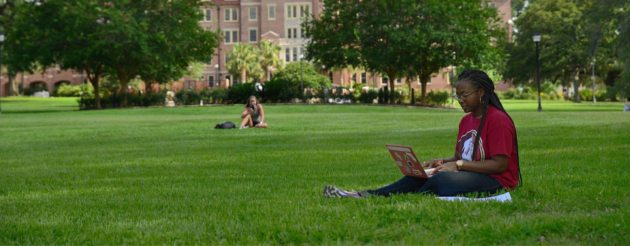 Students sitting in grass on campus with laptop.